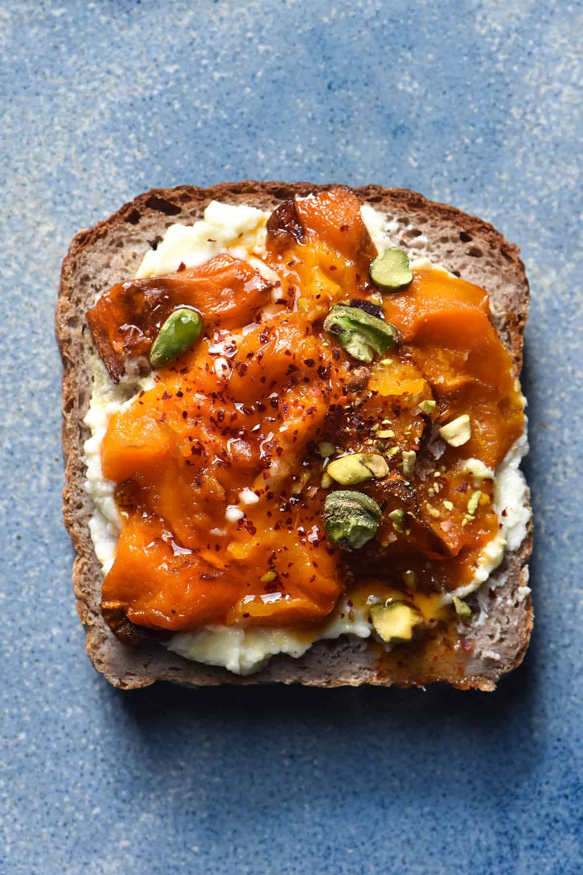 An aerial close up view of a piece of gluten free buckwheat bread on a sky blue ceramic plate. The bread is topped with mascarpone, smashed roasted pumpkin, garlic infused chilli oil and a few chopped pistachios.