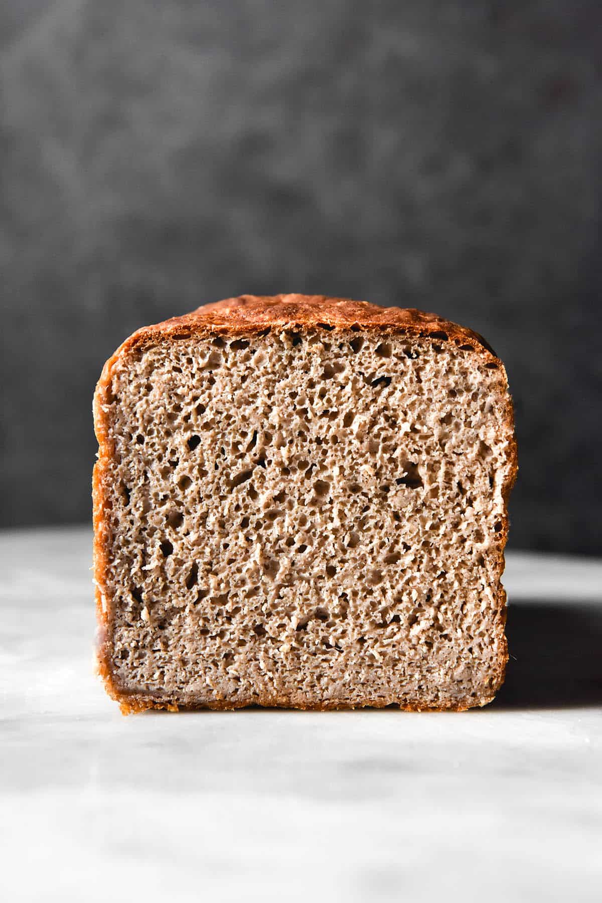A side on view of the crumb of a gluten free buckwheat bread loaf. The bread sits on a white marble table against a dark backdrop