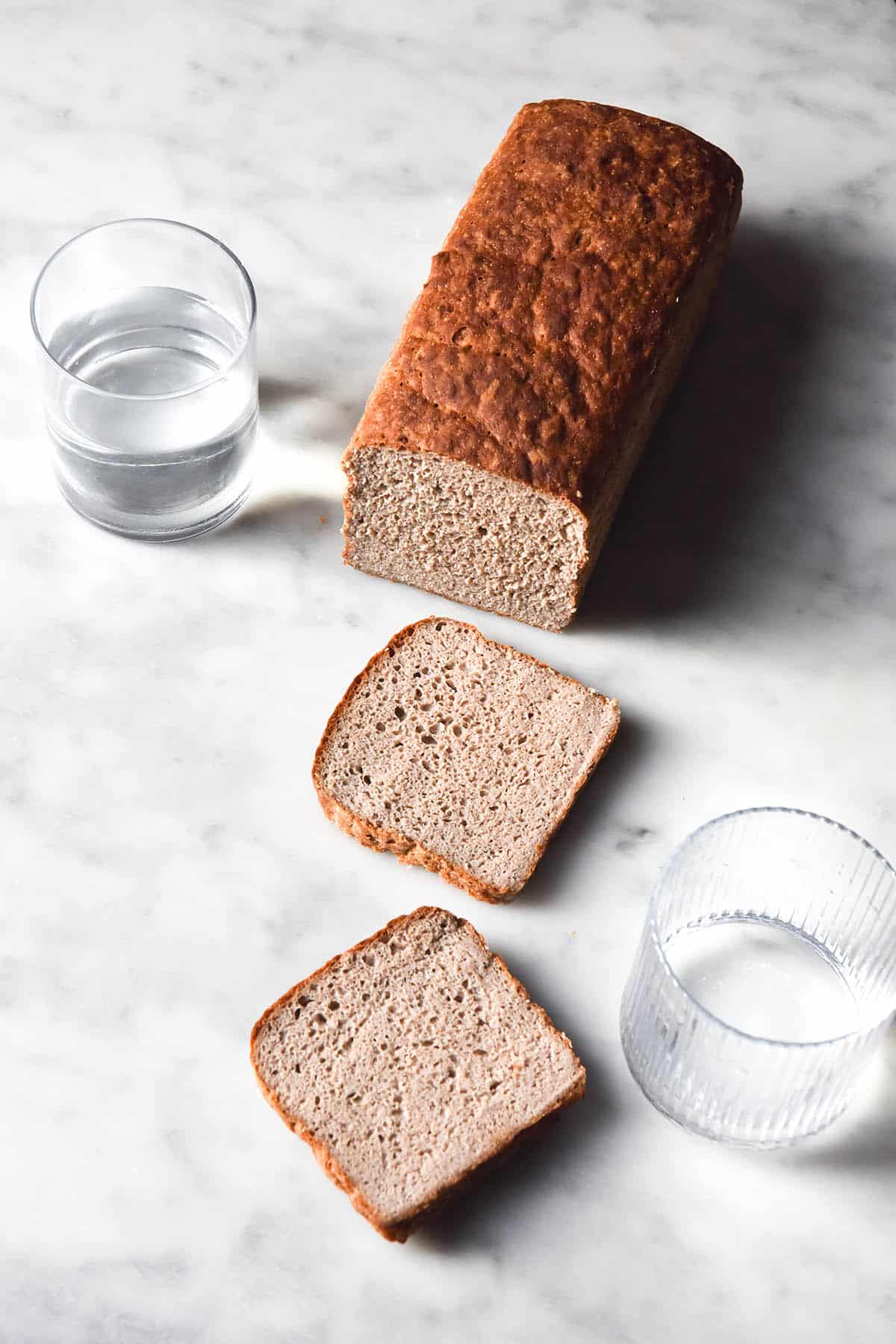 An aerial view of a loaf of gluten free buckwheat bread on a white marble table. The loaf has been sliced, and two slices lay before it on the table. 