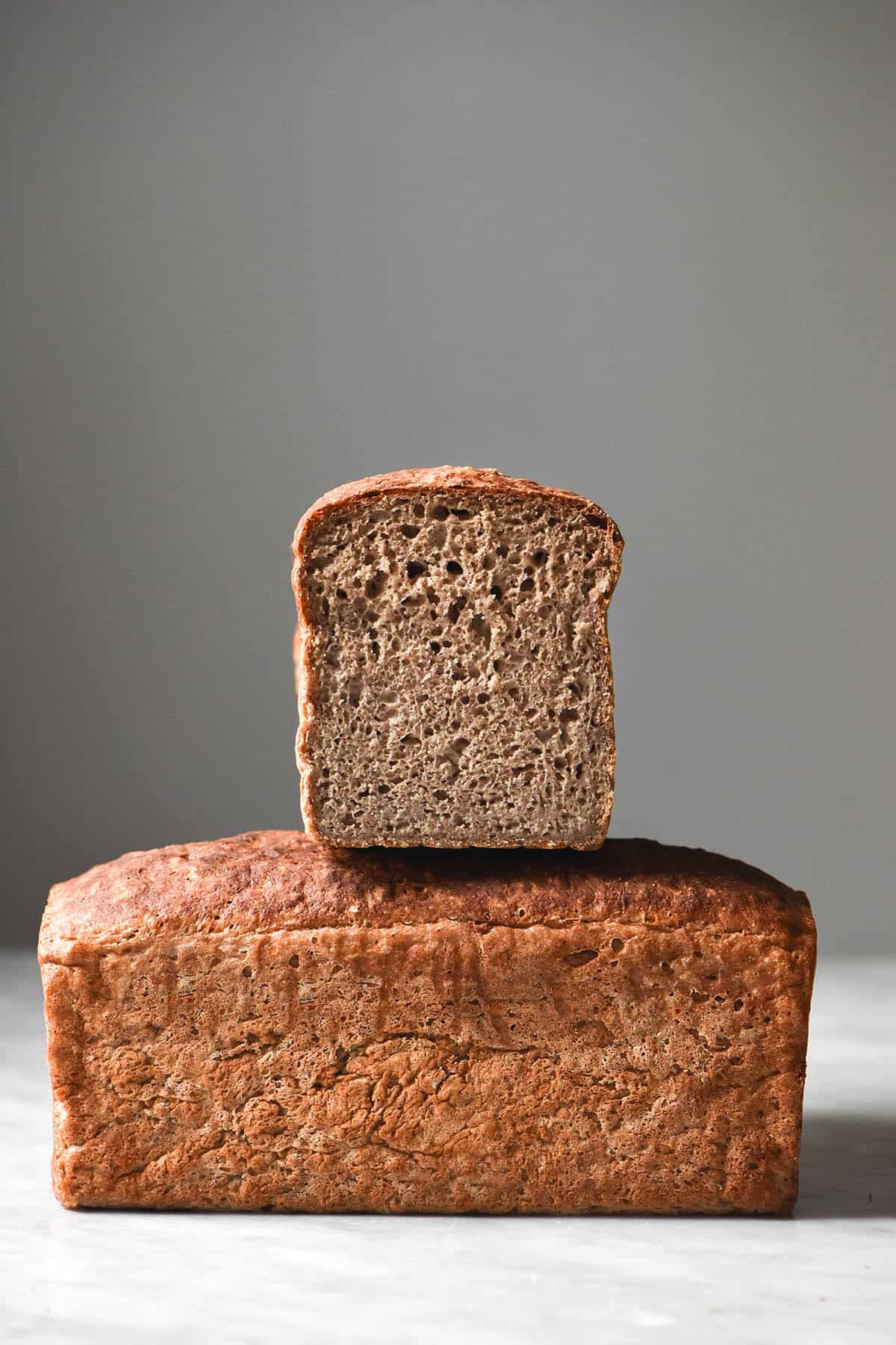 A side on view of a gluten free buckwheat bread loaf on a white marble table. A second loaf sits in the middle of the loaf, facing the camera. The second loaf has been sliced to reveal the inner crumb of the bread. 