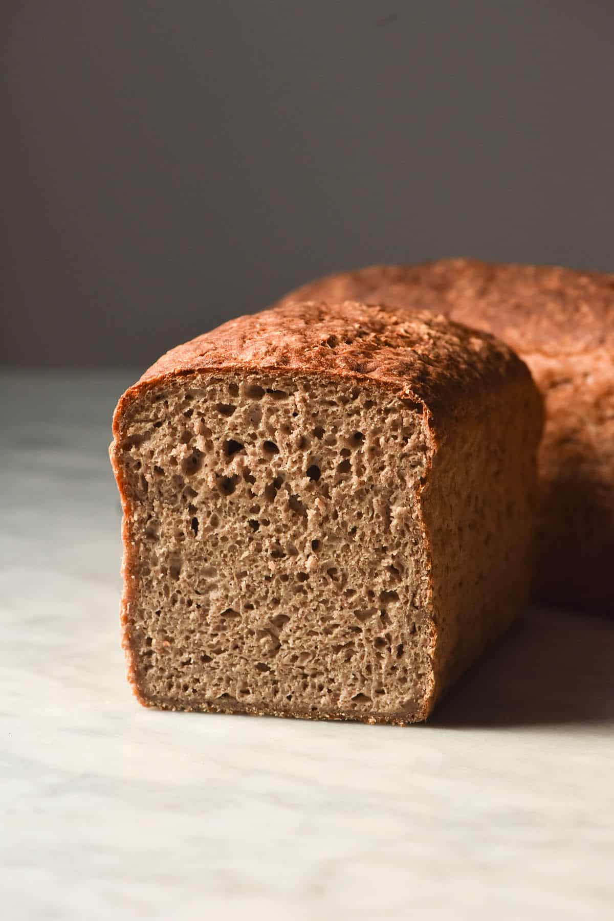 A side on image of a loaf of gluten free buckwheat bread which has been sliced to reveal the crumb inside. Two loaves of the buckwheat bread sit on a white marble table against a light backdrop. 