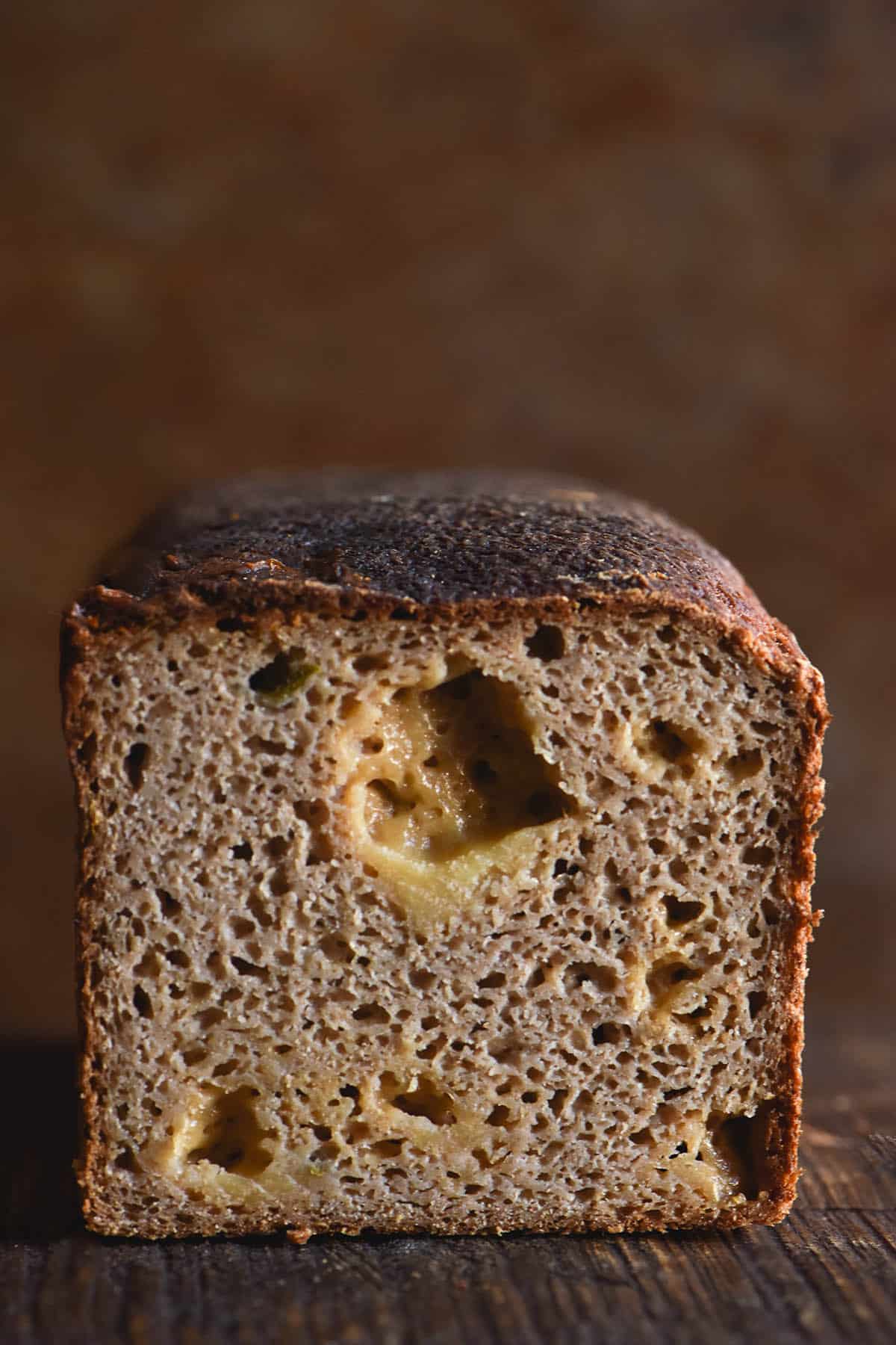 A side on view of the crumb of a gluten free sourdough jalapeno and cheddar loaf. Melty cheddar holes and bits of jalapeno are interspersed with the crumb of the loaf. The bread sits on a wooden table with a dark background