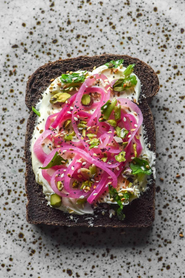 An aerial image of gluten free dark buckwheat bread topped with cream cheese, pickled red onion, chopped pistachios and everything bagel seasoning atop a white speckled ceramic plate
