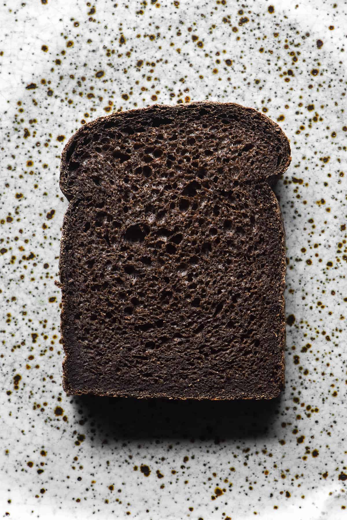 An aerial image of a slice of gluten free buckwheat bread made with dark buckwheat flour. The slice sits atop a white speckled ceramic plate