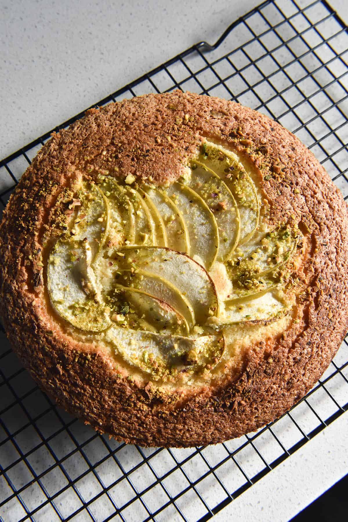 An aerial close up image of a gluten free olive oil cake topped with thinly sliced pear and crumbled pistachios. The cake sits atop a black cooling rack on a white stone bench top. 