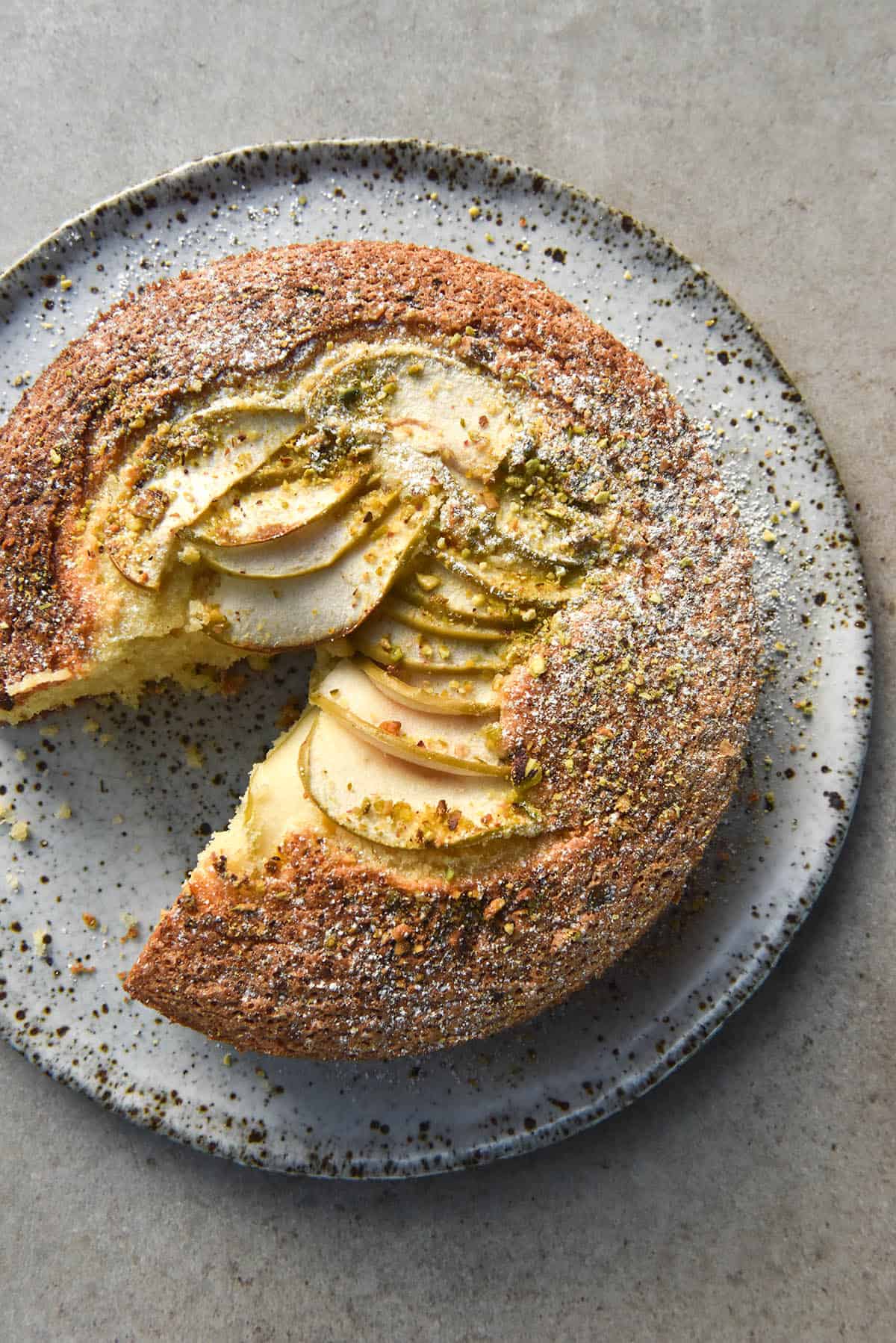 An aerial view of a gluten free olive oil cake topped with thinly sliced apple, chopped pistachios and icing sugar. A slice has been taken from the bottom left side, revealing a white speckled ceramic plate underneath. 