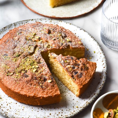 A side on brightly lit image of a gluten free olive oil cake chopped with pistachios and icing sugar. The cake sits atop a white marble table and is surrounded by water glasses, a slice of cake on a white ceramic plate and pinch bowls filled with pistachios