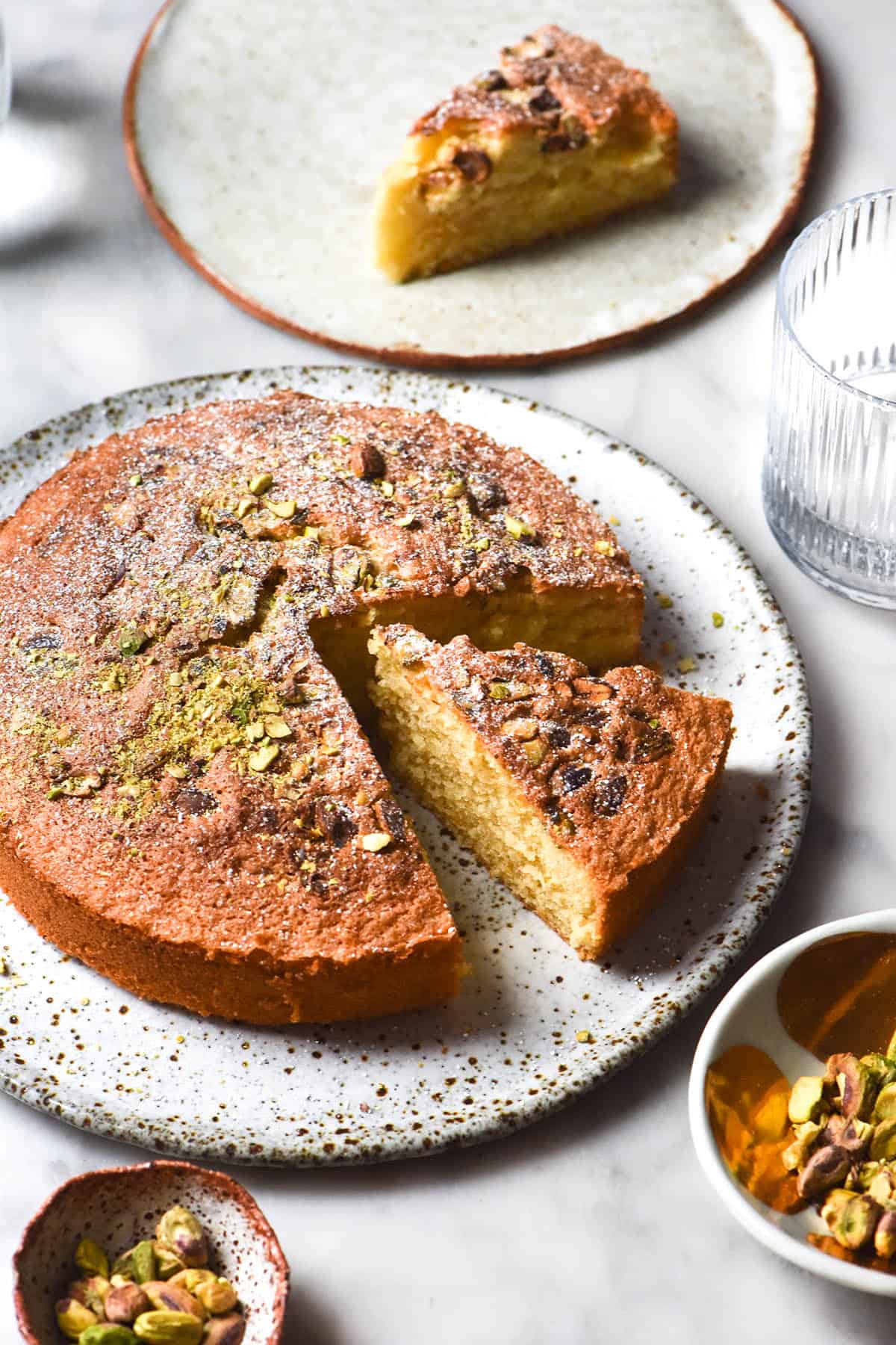 A side on brightly lit image of a gluten free olive oil cake chopped with pistachios and icing sugar. The cake sits atop a white marble table and is surrounded by water glasses, a slice of cake on a white ceramic plate and pinch bowls filled with pistachios
