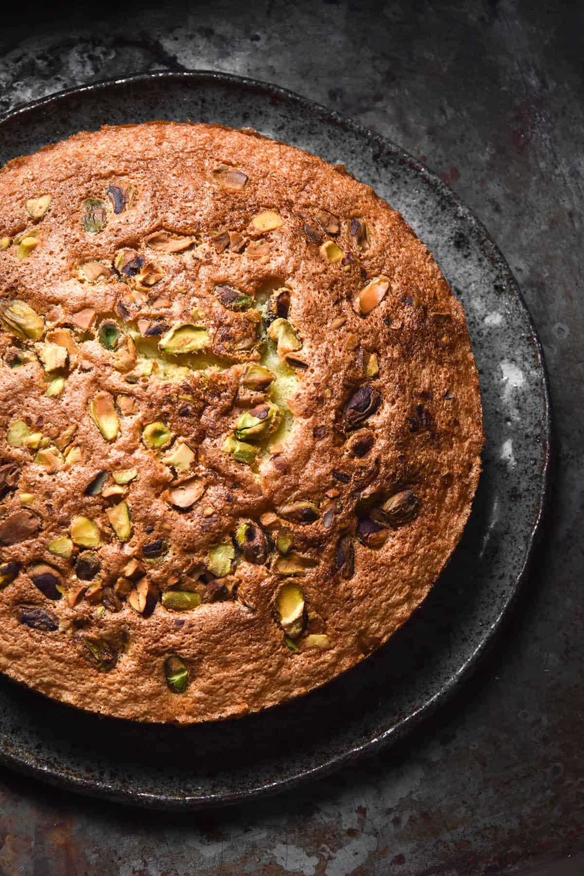 An aerial close up view of the top of a gluten free olive oil cake on a dark grey ceramic plate against a dark steel backdrop. The cake is topped with chopped pistachios and the crumb is golden brown