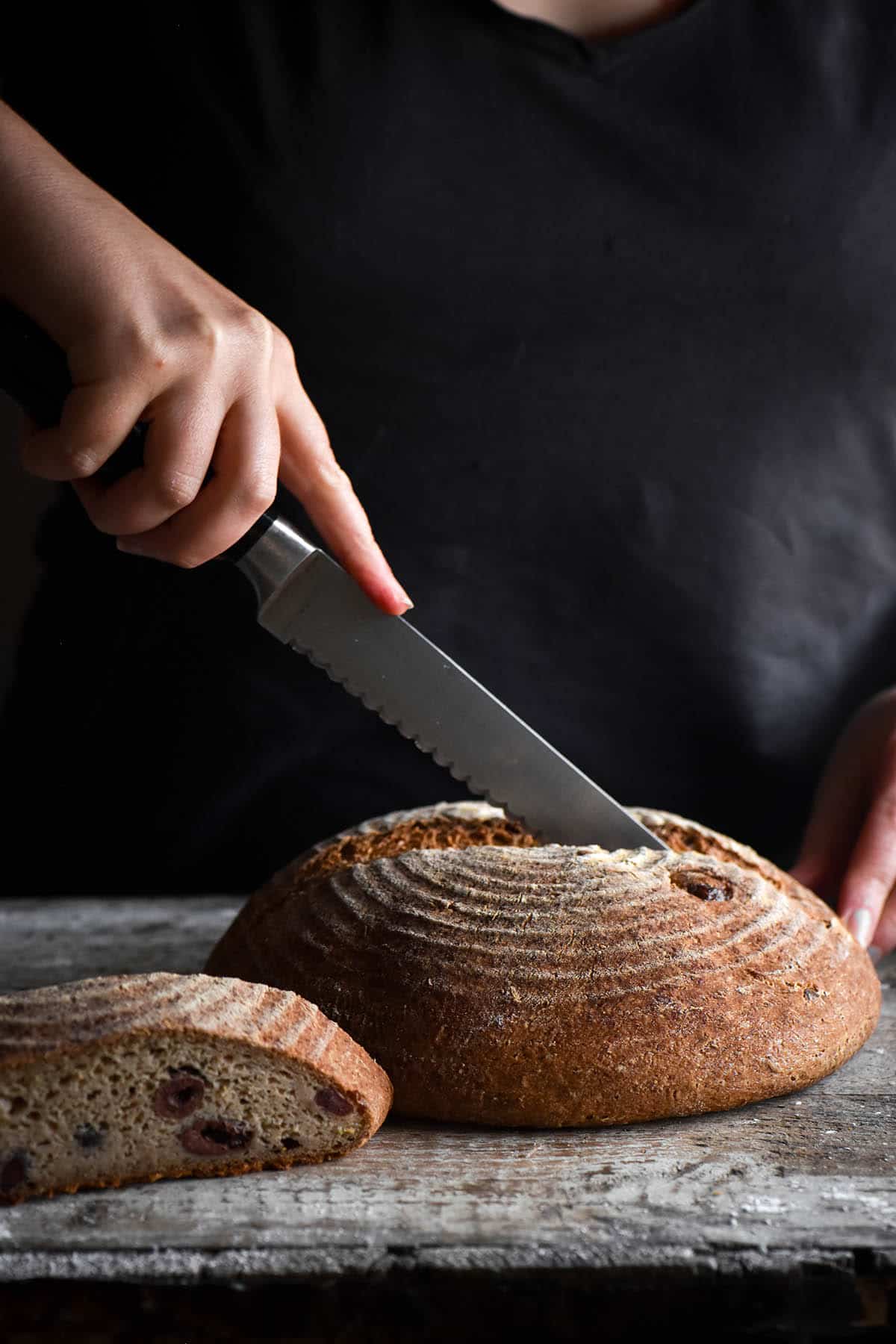 A side on moody image of a gluten free olive and rosemary sourdough boule on a mottled wooden backdrop. A person stands behind the loaf to slice into it it. 
