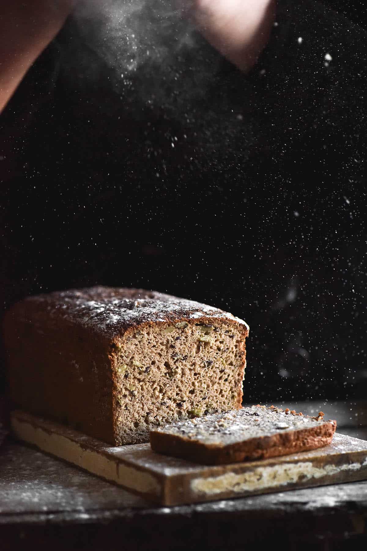 A side on view of a loaf of gluten free seeded bread against a black backdrop. A person stands behind the loaf and sprinkles flour down onto it, which contrasts against the dark backdrop