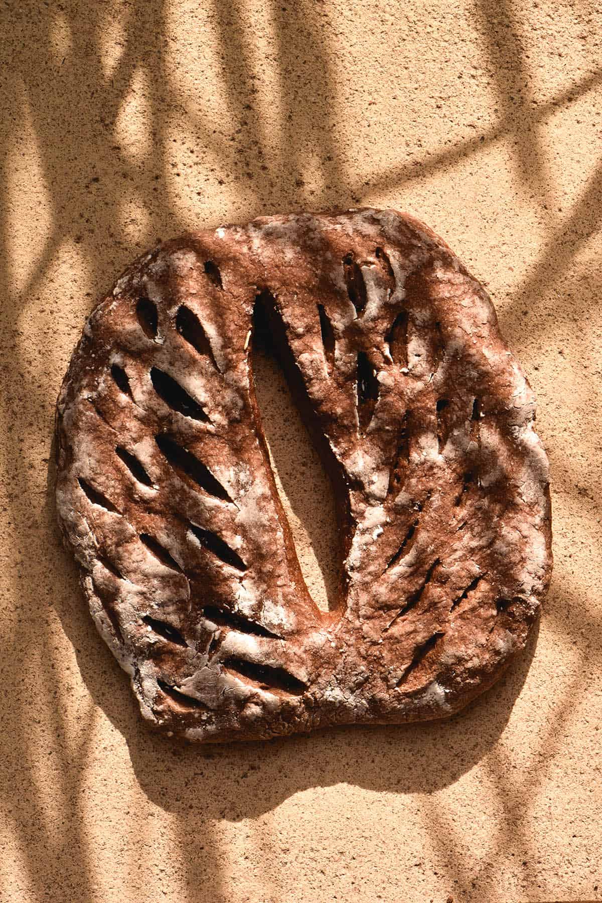 An aerial view of a gluten free fougasse loaf in harsh sunlight. The shadows of palm fronds are reflected back onto the loaf, which sits on a beige stone backdrop. 
