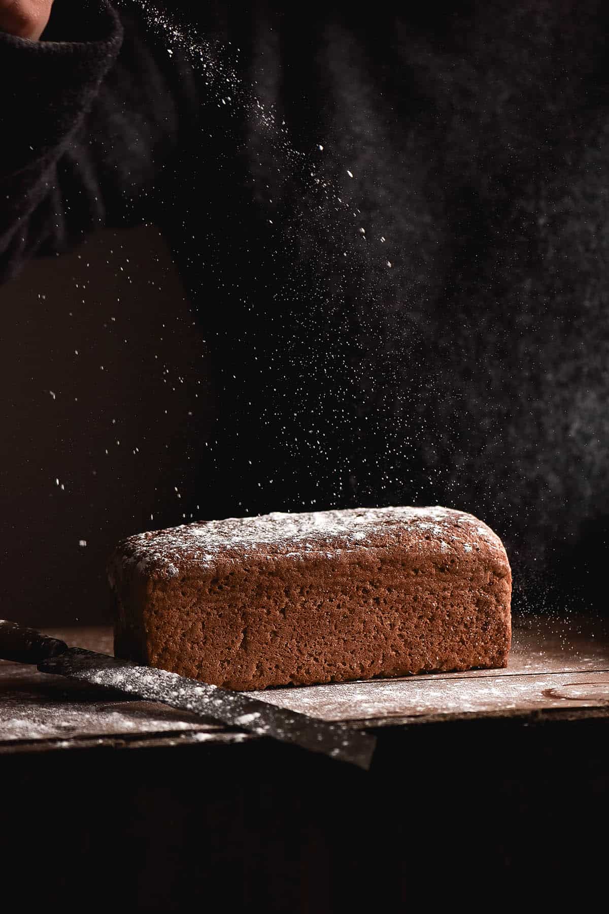 A side on dark image of a loaf of gluten free buckwheat bread. The loaf sits on a wooden table and flour sprinkles down from the top of the image onto the loaf