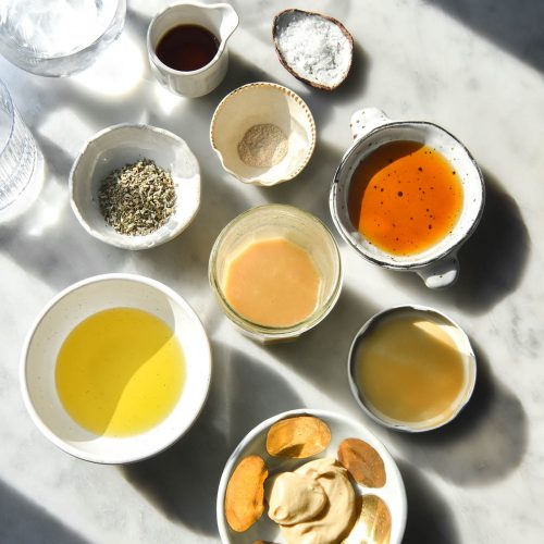 An aerial view of low FODMAP salad dressing and the ingredients used for it arranged on a sunlit white marble table. The ingredients are arranged in small white bowls surrounding the salad dressing in a jar.