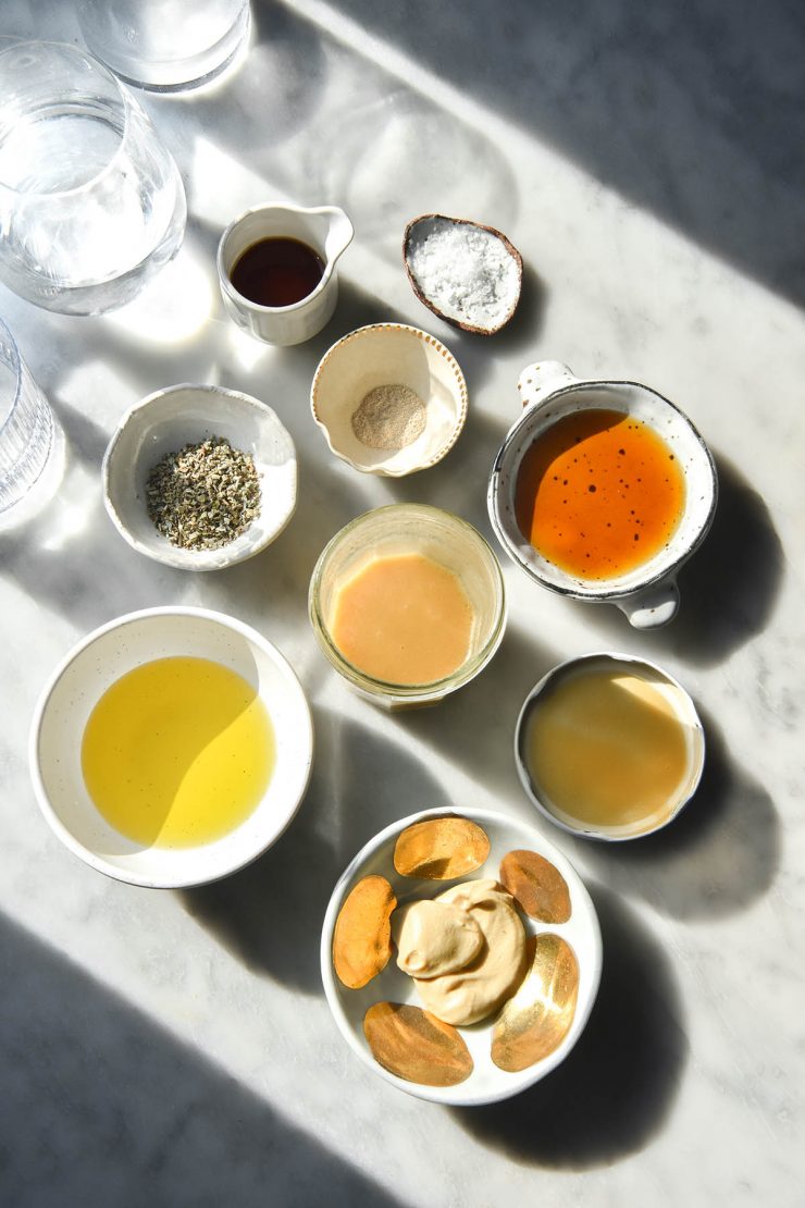 An aerial view of low FODMAP salad dressing and the ingredients used for it arranged on a sunlit white marble table. The ingredients are arranged in small white bowls surrounding the salad dressing in a jar.