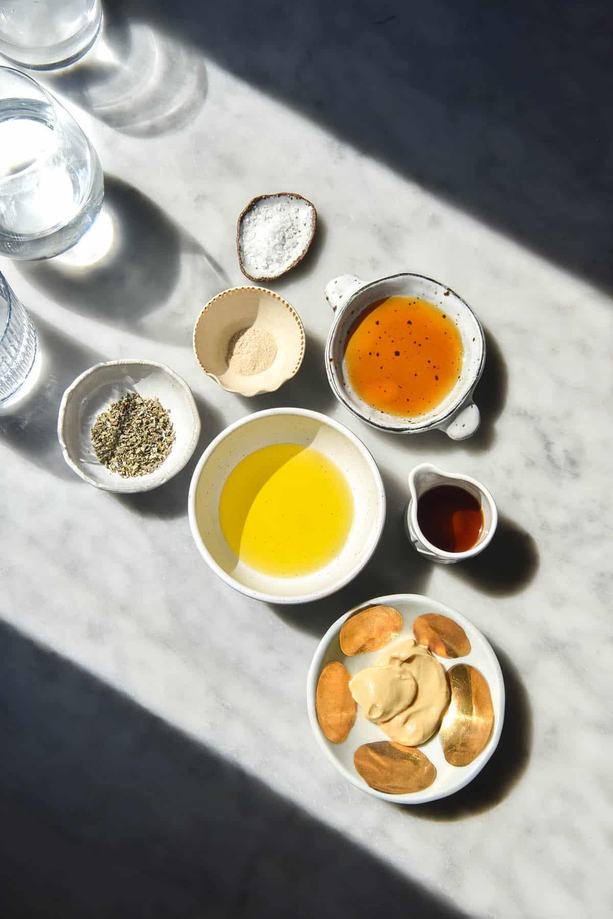 An aerial image of the ingredients used for a low FODMAP salad dressing arranged in small white bowls in a sunlit marble table. Water glasses cast a light and shadow pattern across the image, which is framed by contrasting light