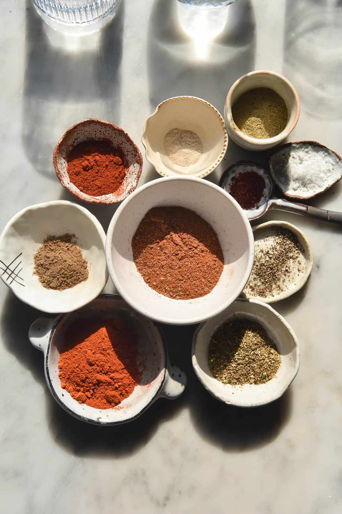 An aerial image of a small white bowl filled with low FODMAP taco seasoning on a white marble table. The bowl is surrounded by other small white bowls filled with the ingredients used to make the taco seasoning. 