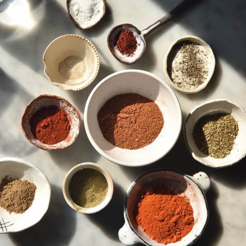 An aerial image of a small white bowl of low FODMAP taco seasoning surrounded by smaller white bowls filled with the ingredients used to make the seasoning. The scene is set on a harshly lit white marble table, and glasses of water create a light and shadow pattern at the top left of the image