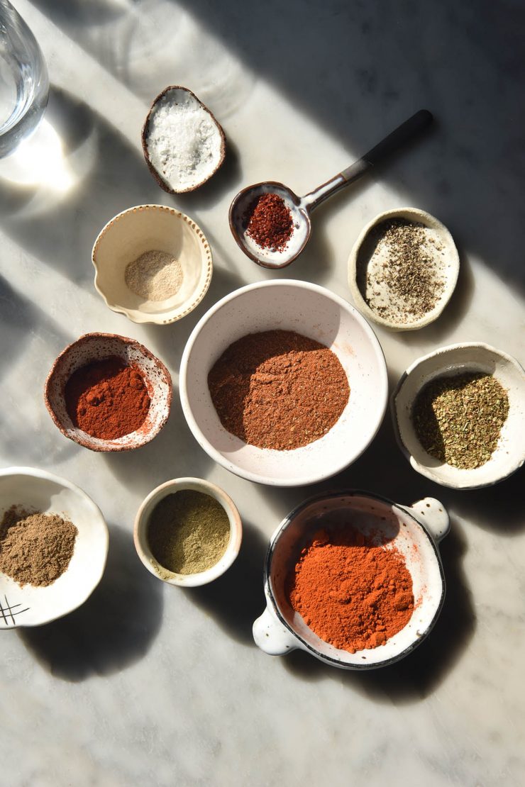 An aerial image of a small white bowl of low FODMAP taco seasoning surrounded by smaller white bowls filled with the ingredients used to make the seasoning. The scene is set on a harshly lit white marble table, and glasses of water create a light and shadow pattern at the top left of the image