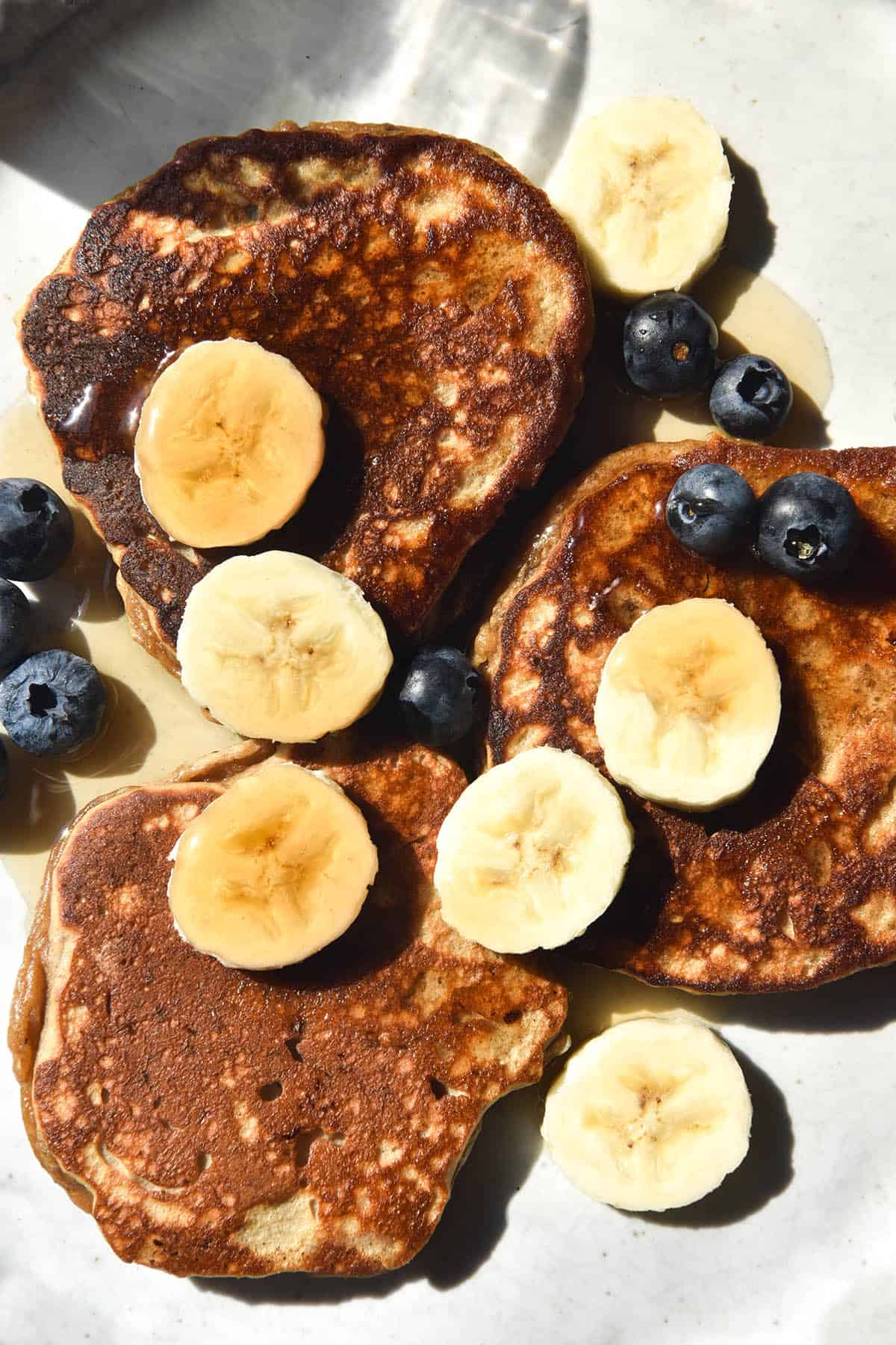 An aerial close up image of protein powder pancakes on a white ceramic plate. The pancakes are topped with sliced banana, blueberries and maple syrup.