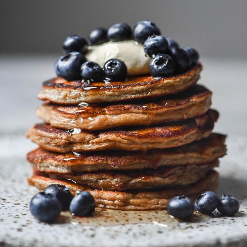 A side on view of a stack of protein powder pancakes topped with mascarpone and berries and drizzled with maple syrup. The pancakes sit on a white speckled ceramic plate atop a white table