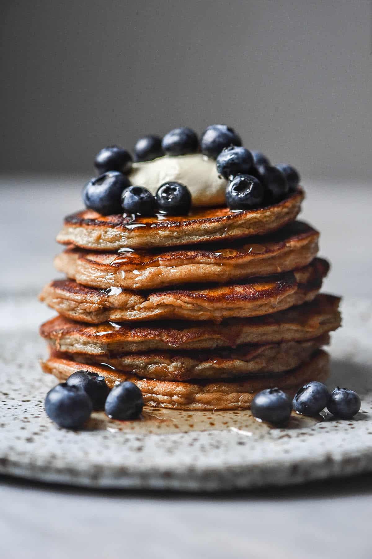 A side on close up view of a stack of protein powder pancakes topped with mascarpone and blueberries and drizzled with maple syrup. The pancakes sit on a white speckled ceramic plate atop a white table. 