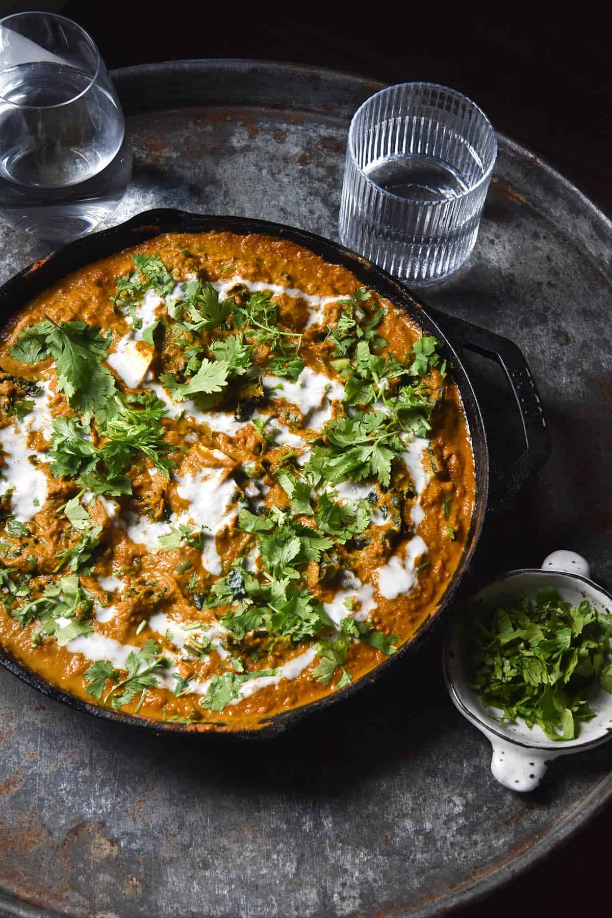 An aerial view of a cast iron skillet filled with SIBO friendly vegan curry. The curry is drizzled with coconut cream and topped with lots of chopped coriander. It sits on a blue steel tray against a black backdrop