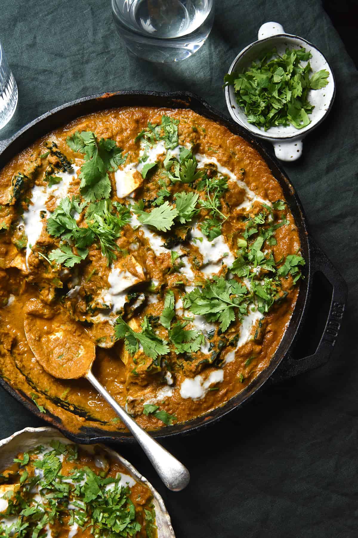 An aerial view of a cast iron skillet filled with SIBO friendly vegan curry. The curry is topped with a drizzle of coconut cream and plenty of coriander. The skillet sits atop an olive green tablecloth and is surrounded by a bowl of curry, water glasses and a pinch bowl with extra coriander