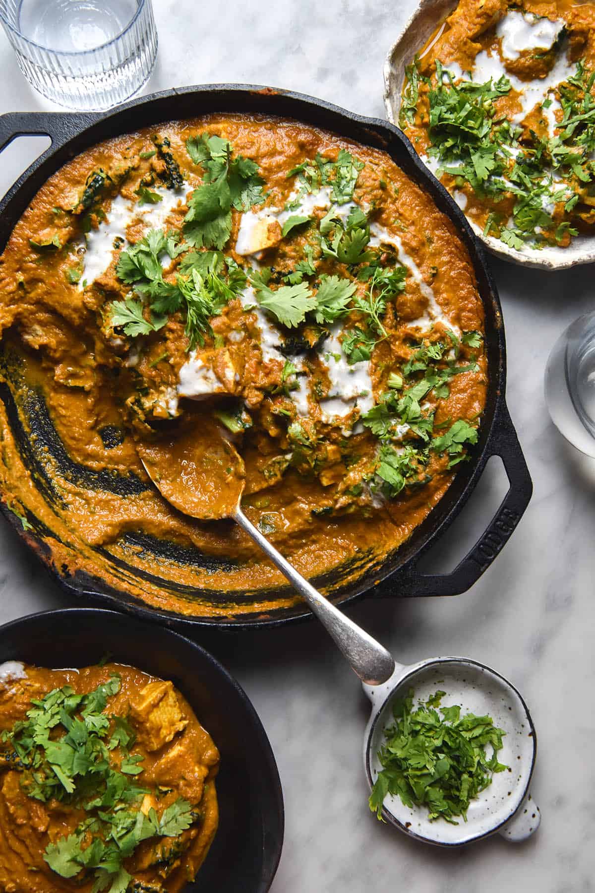 An aerial view of a skillet of SIBO friendly vegan curry on a white marble table. The curry is topped with a drizzle of coconut cream and lots of chopped coriander. A spoon rests on the bottom half of the skillet and an extra bowl of curry sits to the top right
