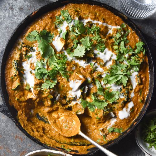 An aerial view of a SIBO friendly vegan curry in a black skillet atop a blue metal backdrop. Water glasses sit to the top of the image, catching the light. A bowl of curry sits in the bottom left of the image