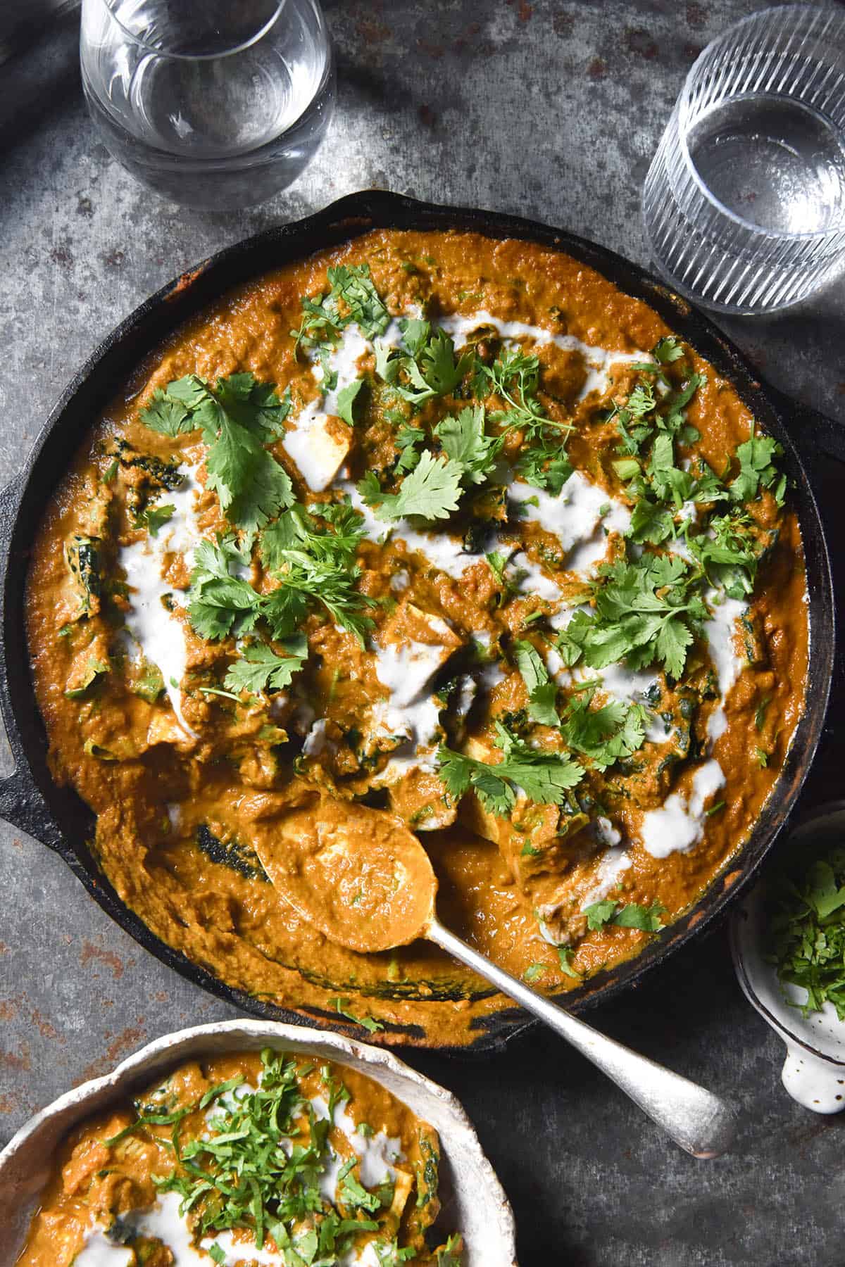 An aerial view of a SIBO friendly vegan curry in a black skillet atop a blue metal backdrop. Water glasses sit to the top of the image, catching the light. A bowl of curry sits in the bottom left of the image