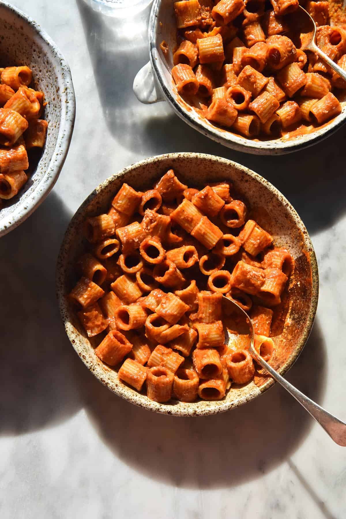 An aerial sunlit view of three bowls on a white marble table filled with tomato mascarpone pasta