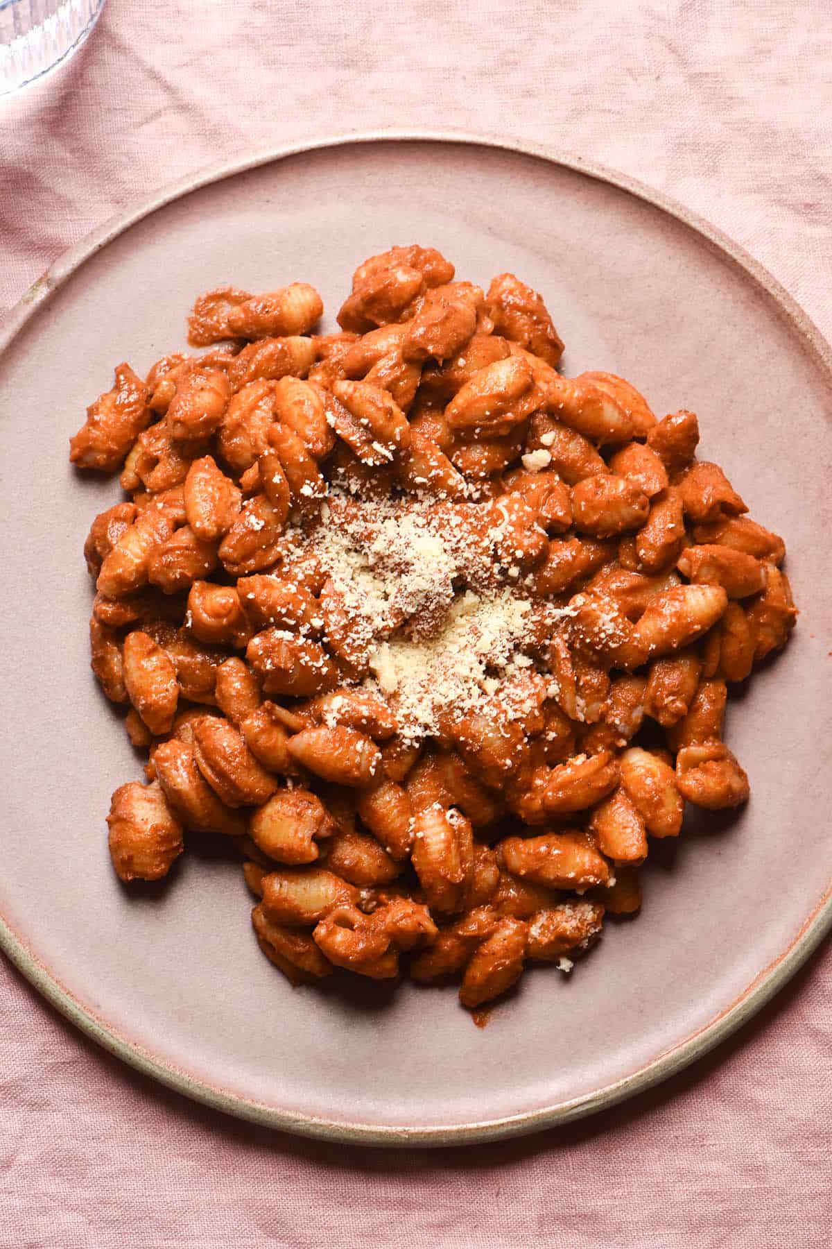 An aerial close up view of a plate of tomato mascarpone pasta topped with parmesan. The pasta shape is a small shell and it sits upon a pale pink ceramic plate which in turn sits on a pale pink linen cloth. 