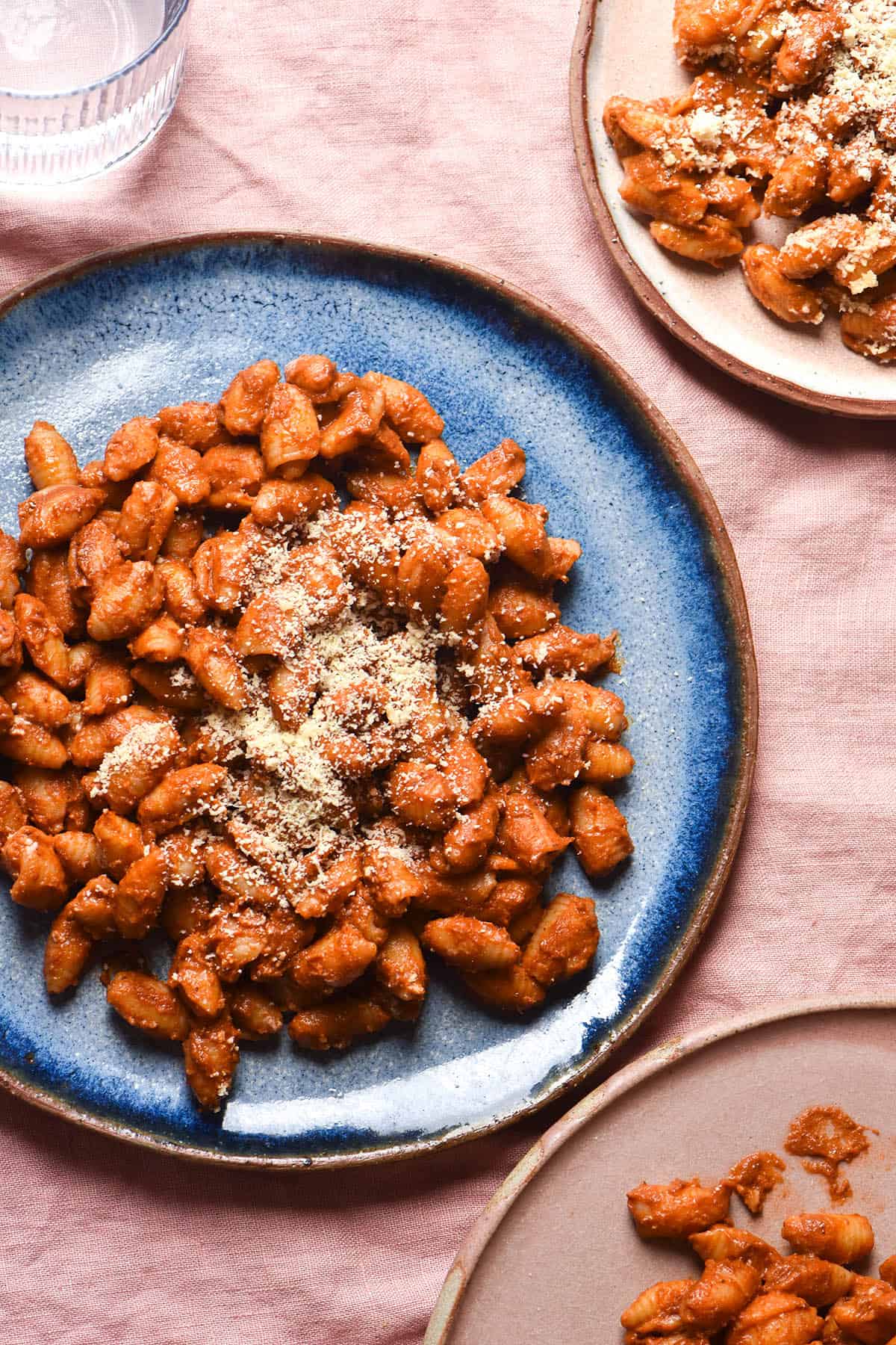 An aerial view of three plates of tomato mascarpone pasta casually arranged on a pale pink linen tablecloth. The central plate is a bright sky blue ceramic plate that contrasts with both the linen and the bright red pasta.