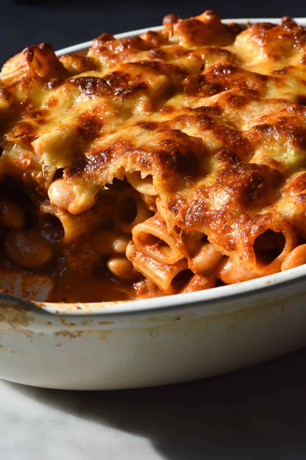 A moody side on image of a tomato mascarpone pasta bake in a white ceramic baking dish. The pasta bake has been cut into, revealing the saucy rigatoni and beans underneath and the melty cheese topping.