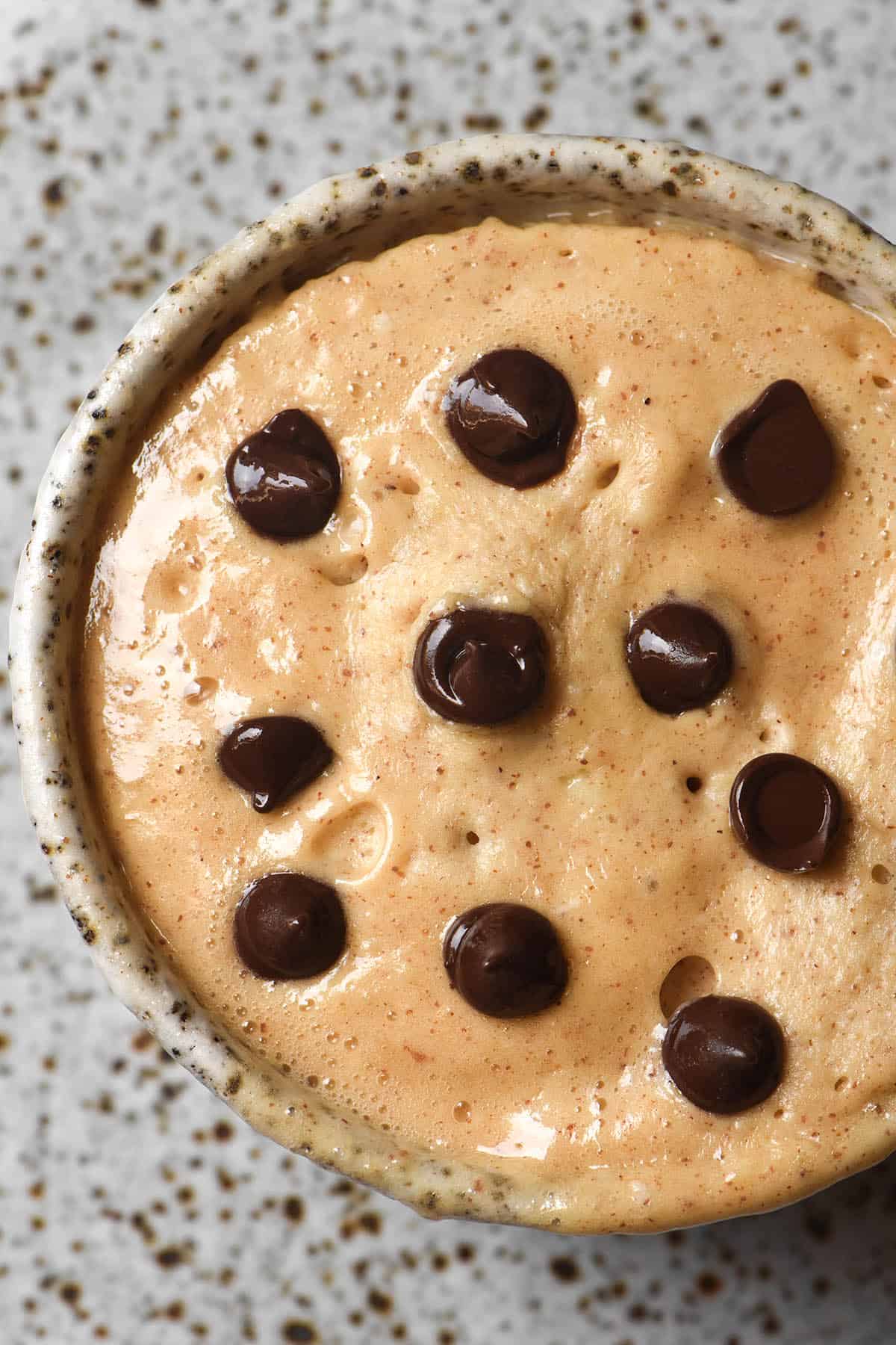 An aerial close up view of a whey protein mug cake topped with chocolate chips. The cake is in a white ceramic speckled mug which sits on a white speckled ceramic plate