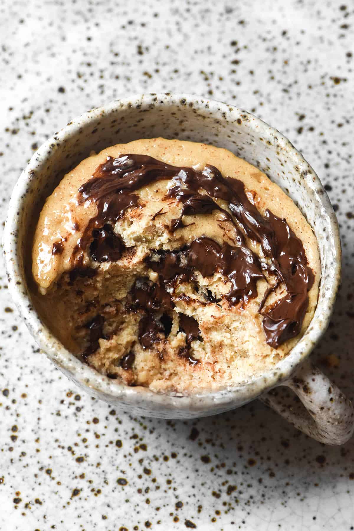 An aerial view of a whey protein mug cake swirled with chocolate chips. A big spoonful has been removed, revealing the mug cake crumb and melted chocolate chips in the centre. The mug sits on a white speckled ceramic plate that acts as the backdrop for the image. 