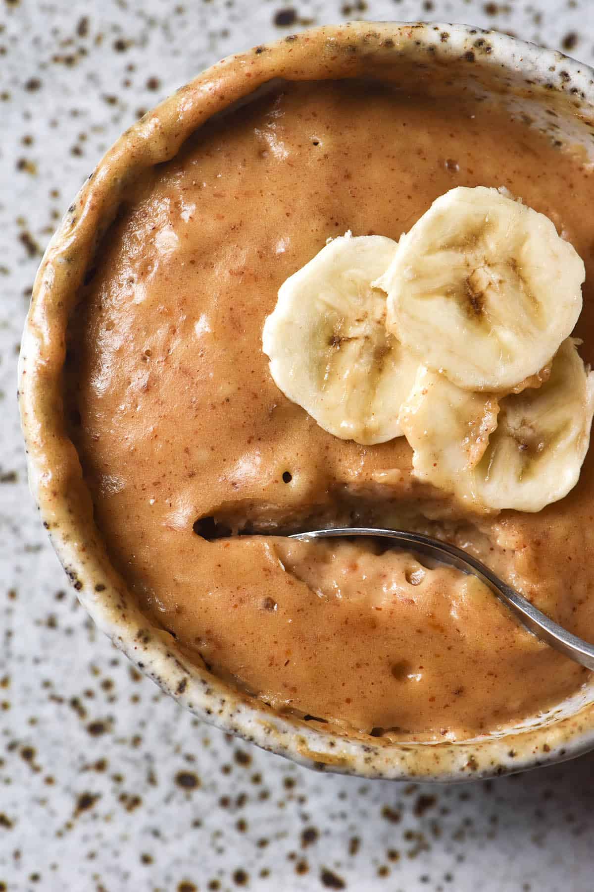 An aerial close up view of a white speckled ceramic mug filled with a protein mug cake. The mug cake is topped with banana slices and a spoon sticks into the cake, revealing the gooey, cakey interior. The mug sits on a white speckled ceramic plate which forms the backdrop