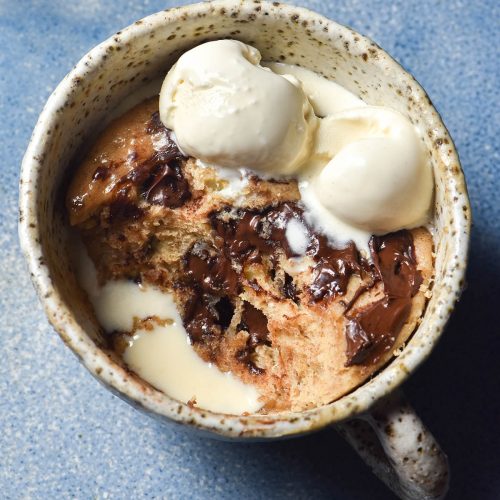 An aerial view of a white speckled mug filled with a whey protein mug cake. The mug cake contains melty chocolate chips and melting vanilla ice cream, which pools together in a hole made by a spoonful of cake being removed. The mug cakes on a bright blue ceramic plate.