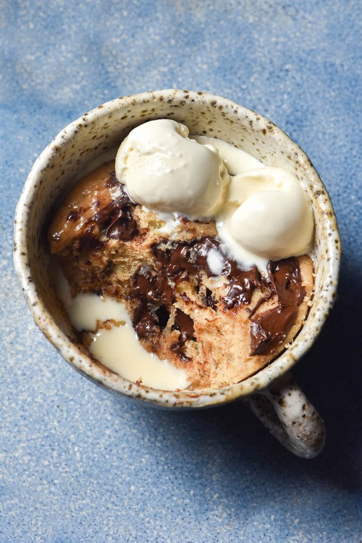 An aerial view of a white speckled mug filled with a whey protein mug cake. The mug cake contains melty chocolate chips and melting vanilla ice cream, which pools together in a hole made by a spoonful of cake being removed. The mug cakes on a bright blue ceramic plate.