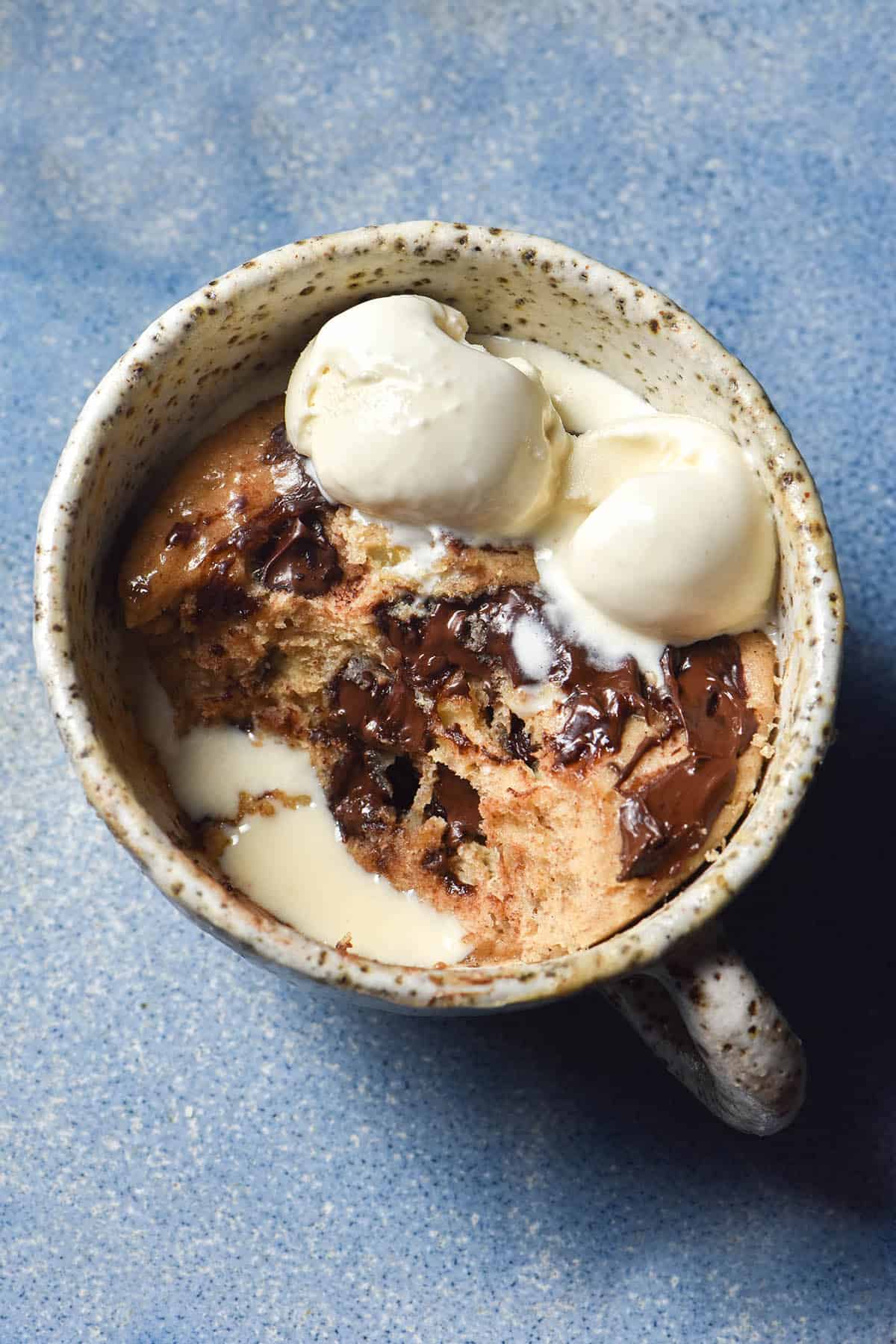 An aerial view of a white speckled mug filled with a whey protein mug cake. The mug cake contains melty chocolate chips and melting vanilla ice cream, which pools together in a hole made by a spoonful of cake being removed. The mug cakes on a bright blue ceramic plate.