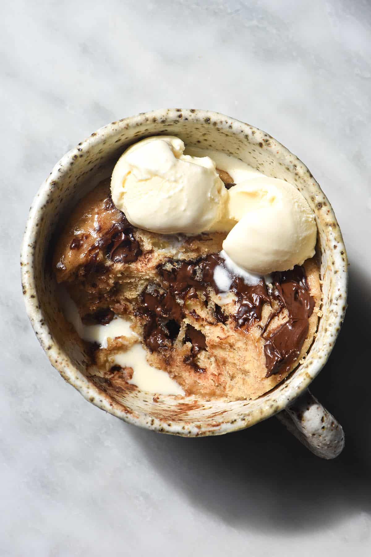 An aerial view of a whey protein mug cake in a white speckled ceramic mug. The mug cake is topped with chocolate chips and melty vanilla ice cream. The mug cake sits atop a white marble table in sunlight. 