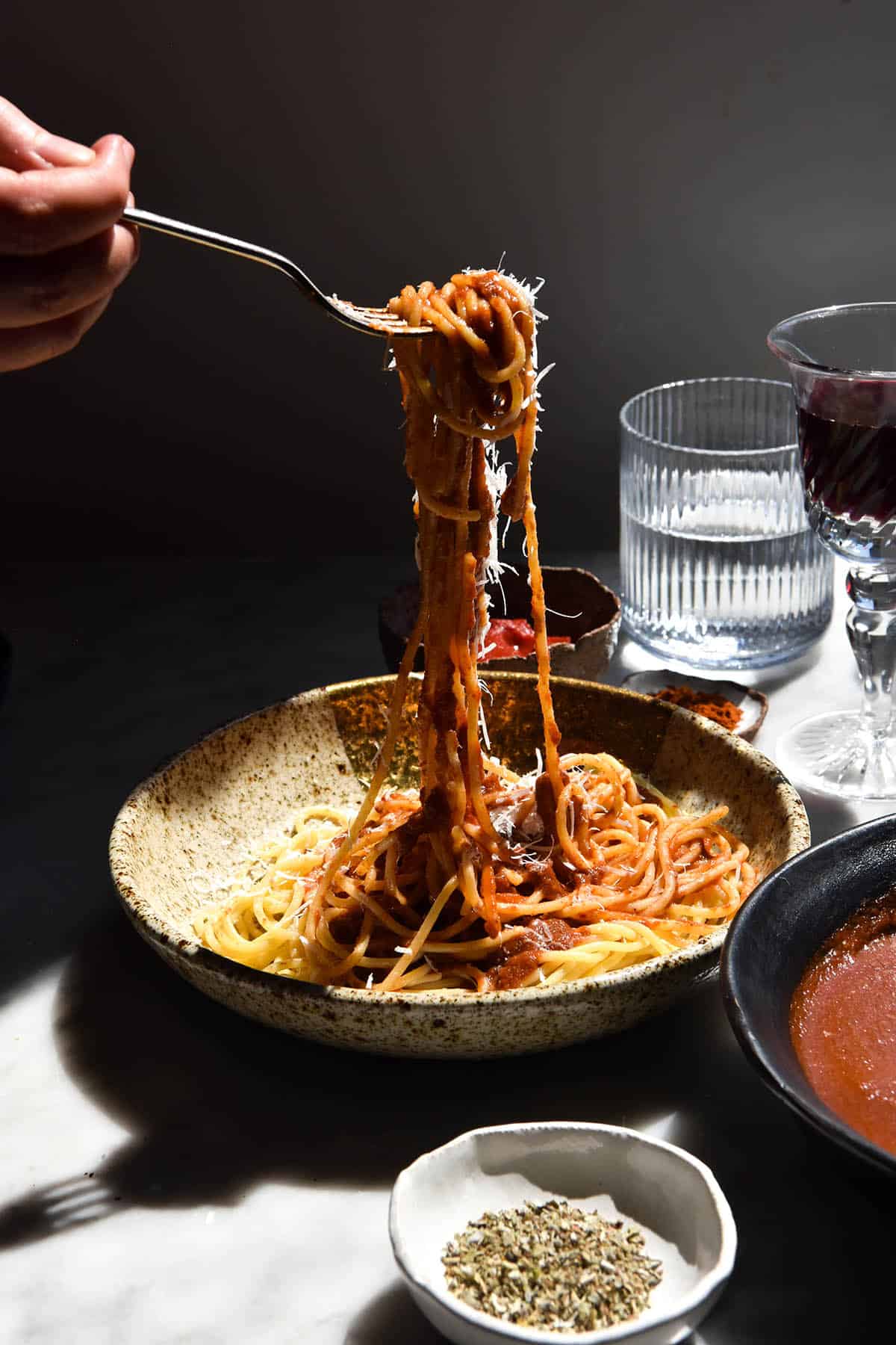 A side on image of a bowl of spaghetti topped with low FODMAP pasta sauce in harsh contrasting light. The bowl sits atop a white marbled table, surrounded by glasses of water, bowls of sauce and pinch bowls of herbs. A hand extends from the left side of the image to hold up a fork full of spaghetti, which is topped with sauce and parmesan