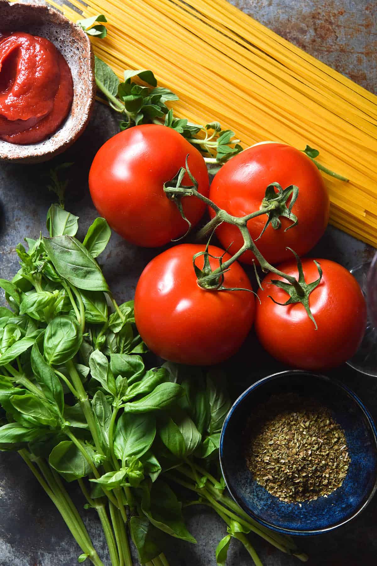 An aerial close up image of vine tomatoes, basil, uncooked spaghetti and pinch bowls of dried herbs and tomato paste arranged on a medium blue steel backdrop