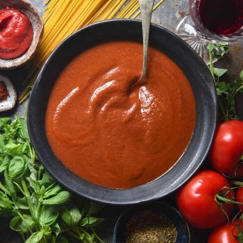 An aerial view of a grey ceramic metal bowl filled with low FODMAP pasta sauce. Surrounding the bowl are pasta ingredients, casually arranged on a medium blue steel backdrop