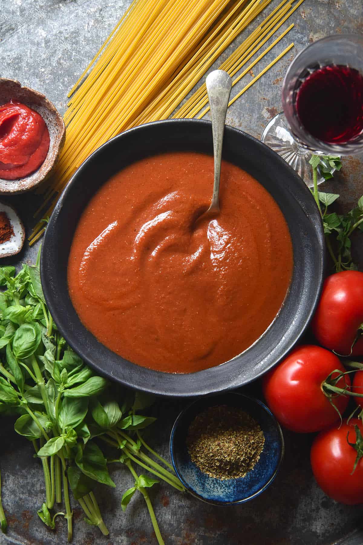 An aerial view of a grey ceramic metal bowl filled with low FODMAP pasta sauce. Surrounding the bowl are pasta ingredients, casually arranged on a medium blue steel backdrop