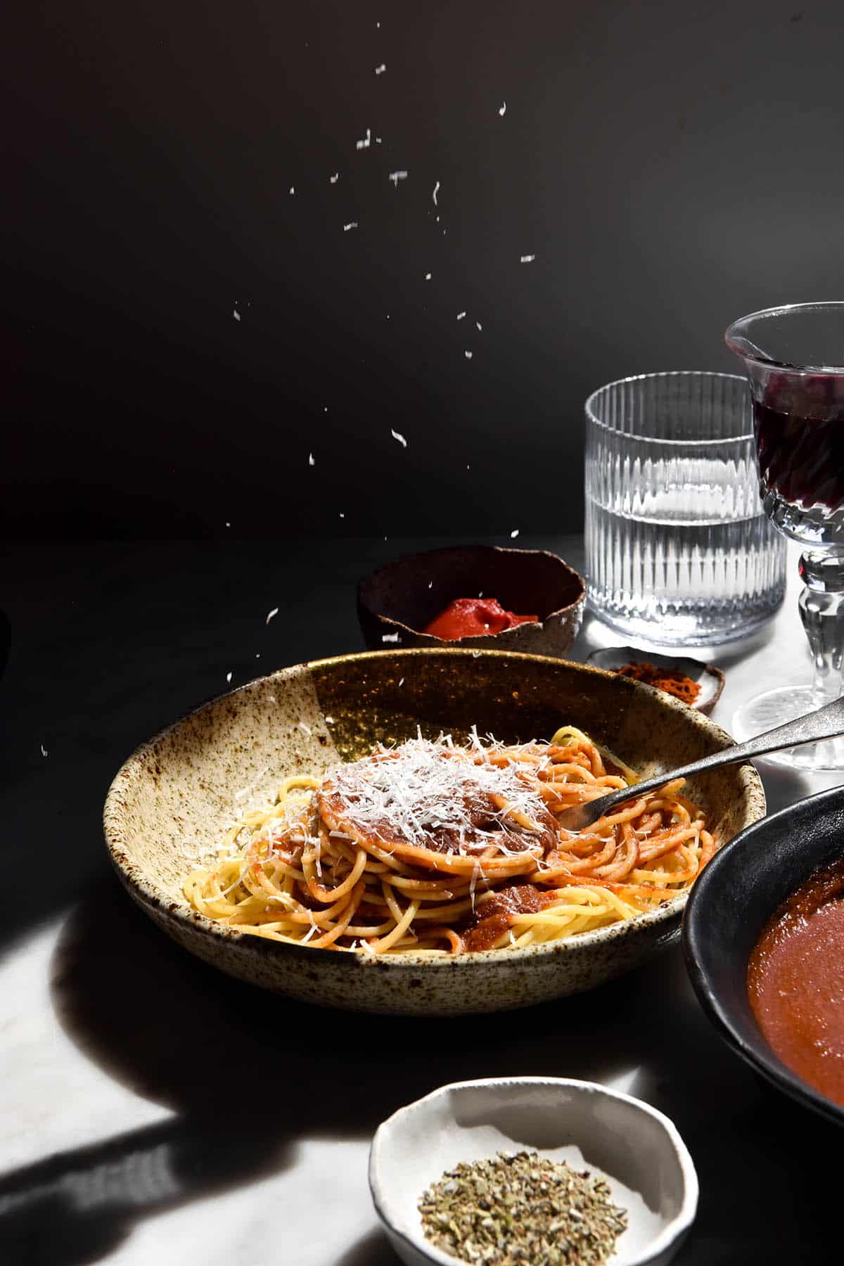 A brightly contrasted side on image of a bowl of spaghetti topped with low FODMAP pasta sauce. The bowl sits on a white marble table surrounded by glasses of wine, water and bowls of the sauce and sauce ingredients. The background is darkened by the contrast of the sun, and flecks of parmesan are grated from the top of the image down onto the pasta. 