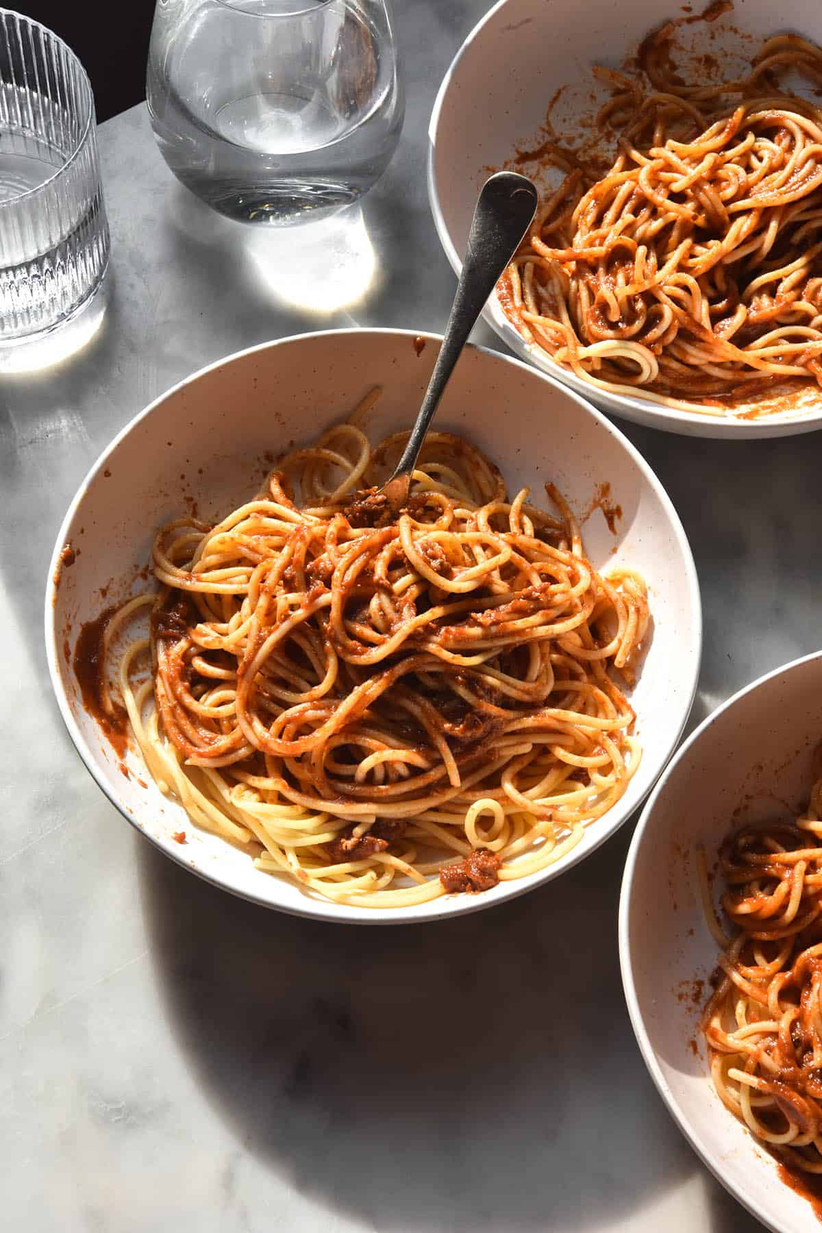 An aerial view of three bowls of spaghetti tossed in low FODMAP pasta sauce atop a white marble table. The table is in contrasting sunlight and the bowls are surrounded by sunlight glasses of water