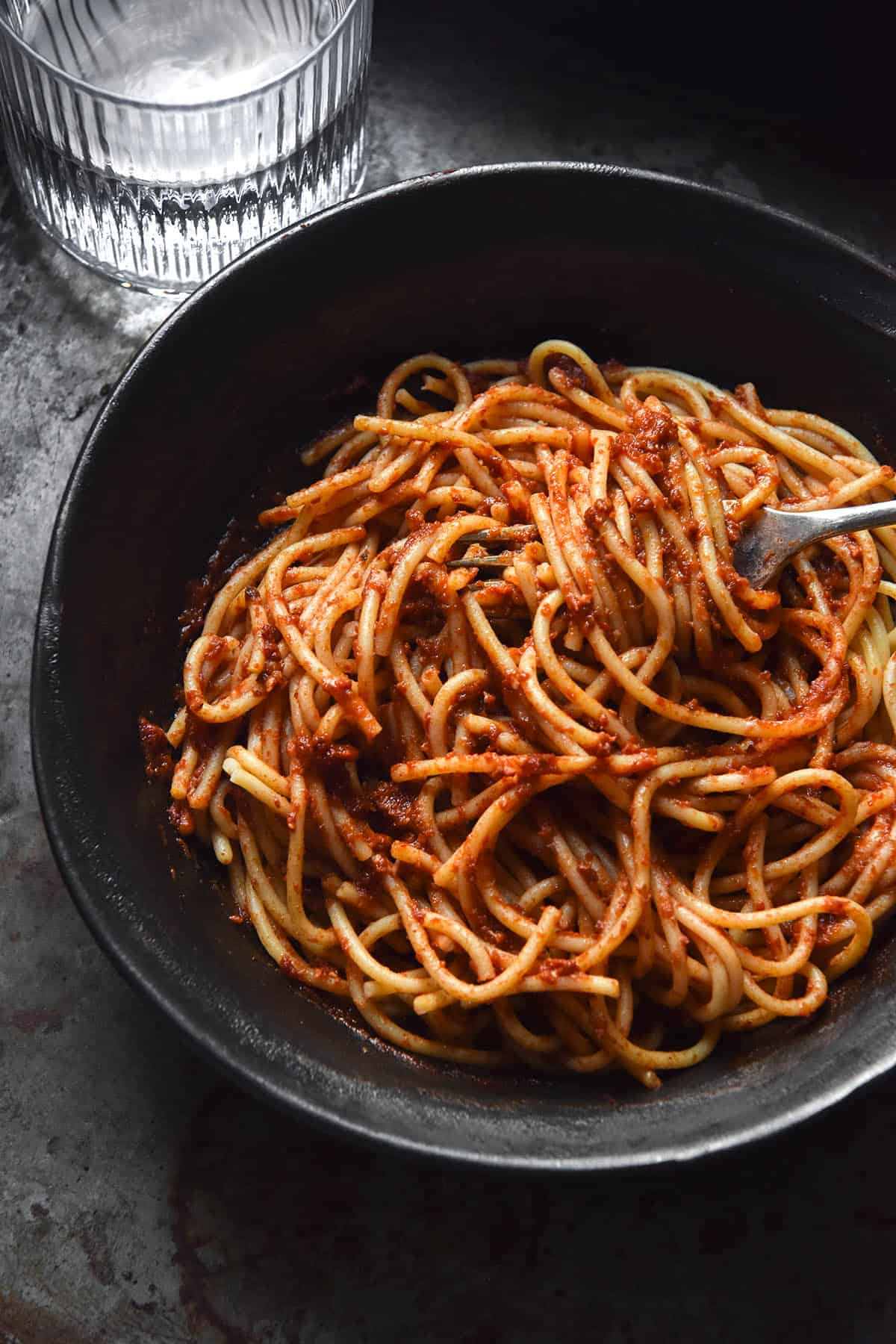 An aerial view of a dark grey ceramic bowl filled with spaghetti tossed in low FODMAP pasta sauce. The bowl sits atop a grey steel background and a fork sticks out of the bowl to the right of the image. A sunlit glass of water sits in the top left corner. 