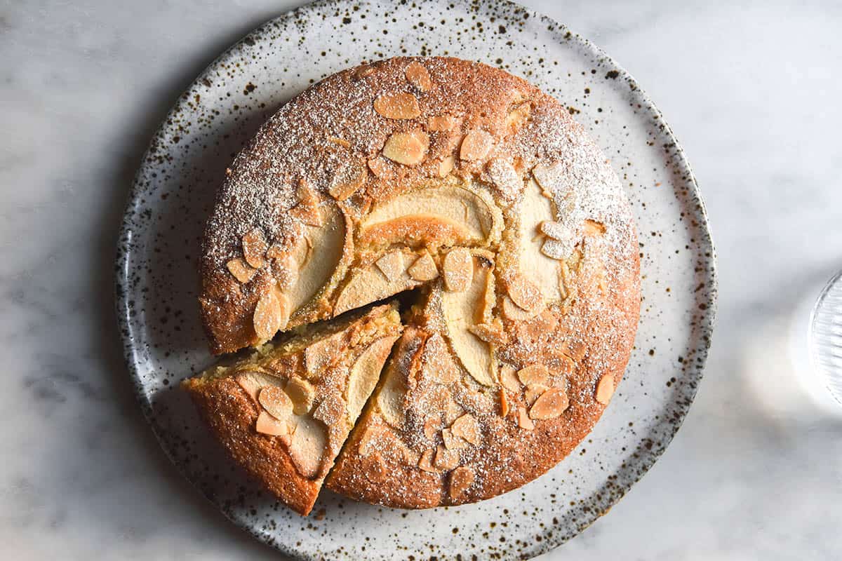 An aerial view of a gluten free apple cake on a white speckled ceramic plate atop a white marble table. The cake is golden brown and topped with slices of apple, sliced almonds and icing sugar